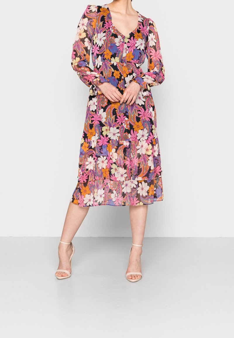 Femme portant une robe midi fleurie colorée à manches longues et col en V, associée à des talons beige clair à bride cheville, debout sur un sol gris.