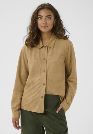 Frau mit lockigem braunem Haar, die ein beige Button-Down-Shirt und dunkle grüne Hosen trägt, steht mit einer Hand in der Tasche vor einem weißen Hintergrund.