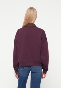 Felpa viola corta con colletto a coste marrone, con spalle scese e polsini lavorati a maglia, abbinata a pantaloni in denim blu.