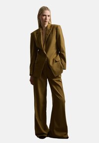 Blazer marrone sartoriale con spalle pronunciate e rever a scollo a V, abbinato a pantaloni a gamba larga. Tessuto liscio con dettagli minimi.