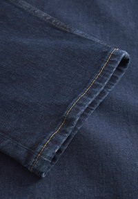 Temno jeans tkanina s prepogibom na robu. Opaženo rumeno šivanje vzdolž roba, z gladko teksturo in subtilnimi barvnimi odstopanji.