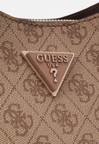 Brun mönstrad tyghandväska med en triangulär metallisk Guess-logotyp som har ingraverad text och ett frågetecken.