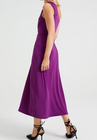 Fioletowa sukienka bez rękawów z dopasowanym gorsetem i rozkloszowanym dołem. Posiada odkryte plecy i gładką, fakturowaną tkaninę. Do kompletu czarne buty na wysokim obcasie.