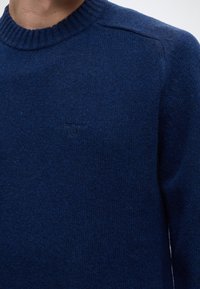 Gros plan d'une personne portant un pull en maille bleu foncé avec un col rond côtelé et un logo brodé discret sur la poitrine.