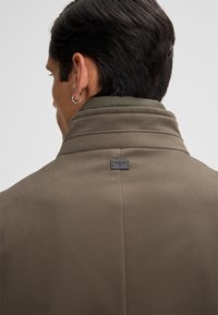 Bruine jas met een hoge kraag, voorzien van een contrasterende groene binnenvoering en een klein zwart logo-label nabij de kraag. Gladde textuur.