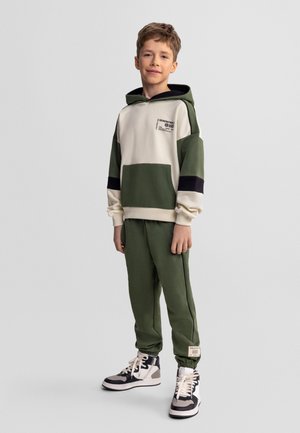 Chłopiec stojący, ubrany w zielono-kremową bluzę z kapturem i dopasowane spodnie dresowe oraz czarno-białe wysokie trampki.