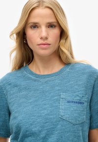Teal bomulds T-shirt med korte ærmer, rund hals, frontlomme med broderet "SUPERDRY" i mørkeblå. Blødt, tekstureret stof.