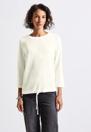 Haut texturé crème clair avec des manches trois-quarts et un cordon de serrage en bas. Associé à un pantalon gris foncé, doté d'un col rond.