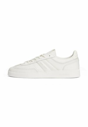 White leather sneaker with textured upper, logo accents, rounded toe, flat laces, and a rubber sole featuring horizontal grooves.