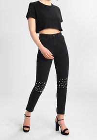 T-shirt nera cropped, jeans skinny neri con decorazioni di perle sulle caviglie e sandali neri con tacco alto. Tessuto liscio, design aderente.