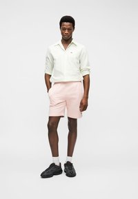Jeune homme debout portant une chemise vert clair boutonnée, un short rose, des chaussettes blanches et des baskets noires sur un fond blanc uni.