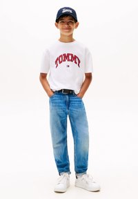 Hvit bomull t-skjorte med rød "TOMMY" tekst, kombinert med blå denimjeans, svart belte, og hvite sneakers. Marineblå cap med logo.