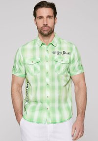 Camisa de manga corta a cuadros verdes que presenta dos bolsillos en el pecho, detalles de botones y un logo en la parte frontal. Tejido ligero con una textura sutil.