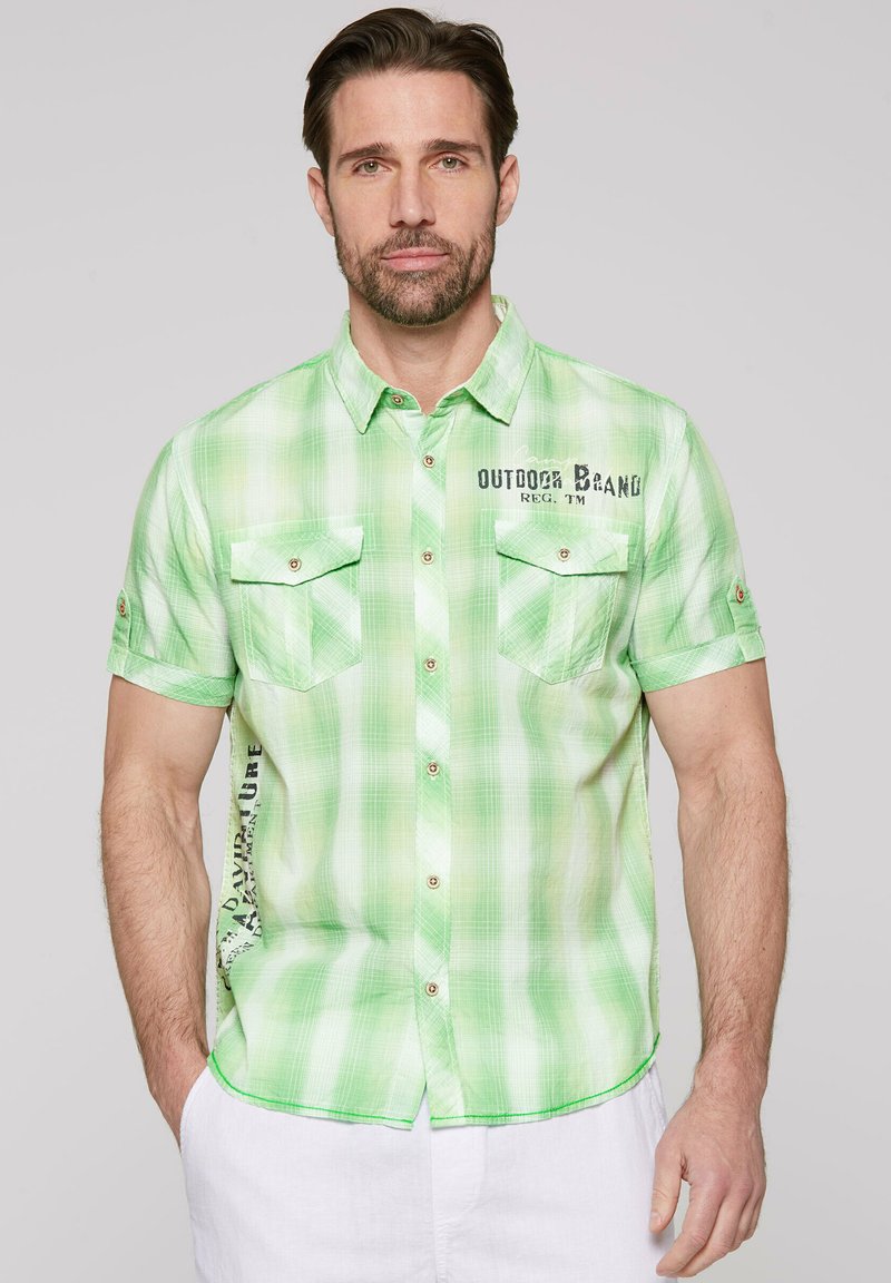 Camisa de manga corta a cuadros verdes que presenta dos bolsillos en el pecho, detalles de botones y un logo en la parte frontal. Tejido ligero con una textura sutil.