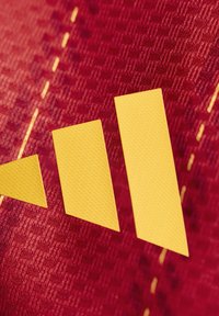 Gele geometrische vormen op een getextureerde rode achtergrond met zichtbare stiksels; vormen omvatten twee grotere diamanten en een kleinere driehoek.