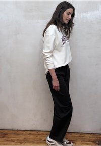 Sweat-shirt blanc avec motif "BOSTON", pantalons noirs à jambes larges et baskets multicolores. Textures lisses, coupe décontractée. Fond en parquet en bois.