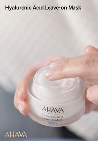 AHAVA Hyaluronsyrabaserad leave-on-mask i en klar, rund burk. Tjock, vit krämig konsistens med en slät yta, hålls med fingrarna ovanför burken.