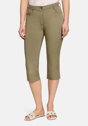 Kvinde iført olivengrønne capribukser og hvide slides, stående foran en neutral baggrund.