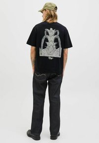 Camiseta de algodón negra con un gran gráfico plateado en la parte posterior. Combinada con jeans oscuros y zapatos negros, presenta un corte relajado y mangas cortas.