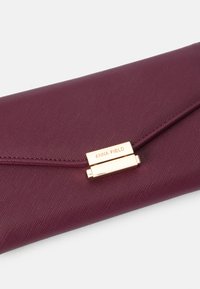Burgundin värinen clutch, joka on valmistettu teksturoidusta kankaasta. Siinä on kultainen metallilukko, jossa on brändin nimi "ANNA FIELD". Kolmionmuotoinen läppädesign.