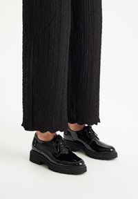 Zapatos de charol negros con puntera redondeada, diseño con cordones y suela gruesa y texturada, llevados con pantalones de tiro ancho negros con textura.