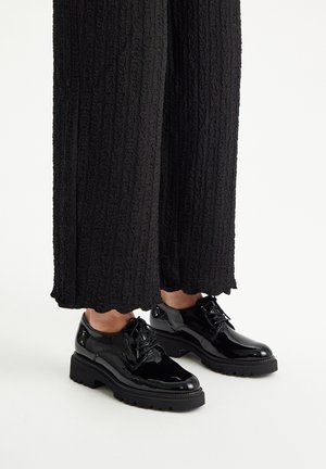 Scarpe in vernice nera con punta arrotondata, design con lacci e suola spessa e testurizzata, indossate con pantaloni larghi neri a trama.
