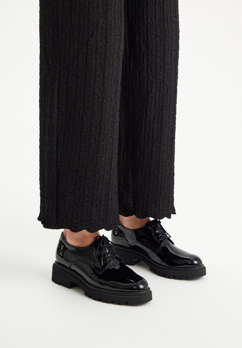 Zapatos de charol negros con puntera redondeada, diseño con cordones y suela gruesa y texturada, llevados con pantalones de tiro ancho negros con textura.