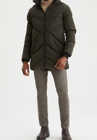 Mørkegrønne puffervest med chevronmønster, frontlynlås og hætte. Båret over en lys brun turtleneck og grå teksturerede bukser, parret med brune støvler.