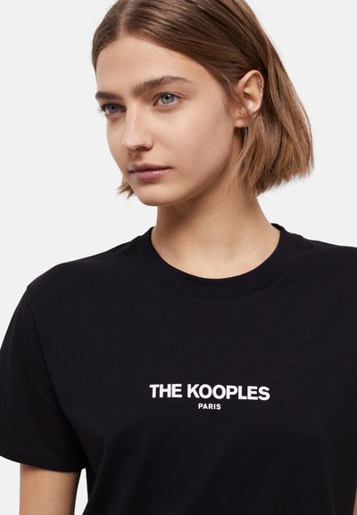 Melnā kokvilnas T-kreklā ar apaļu kakla izgriezumu, īsām piedurknēm un baltu uzrakstu "THE KOOPLES PARIS" priekšpusē.