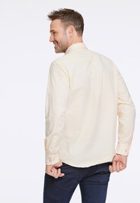 Långärmad, ljusgul skjorta med standardkrage, knapprör och bakveck, bärs med mörka jeans mot en neutral bakgrund.