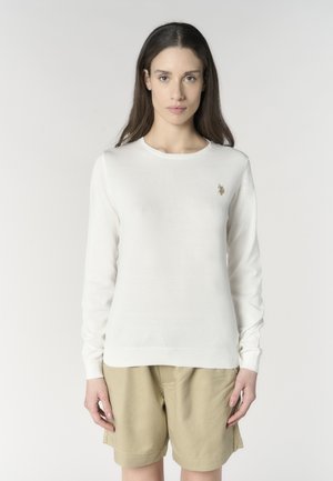 Donna con lunghi capelli castani che indossa un maglione bianco a maniche lunghe e pantaloncini beige, in piedi davanti a uno sfondo chiaro e semplice.