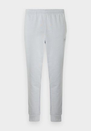 Pantalones deportivos gris claro hechos de material suave, con cinturilla elástica, piernas estrechas y un pequeño parche con logo en el lateral.