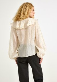 Lys beige bluse med flæsekrave, lange pufærmer og sort prikket mønster. Texturede stof med en løs pasform, parret med mørke bukser.