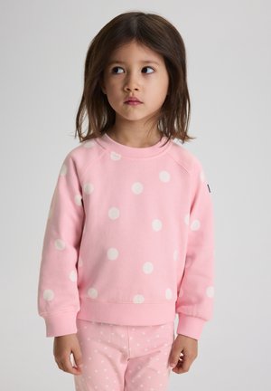 Jong meisje met schouderlang bruin haar, gekleed in een roze sweatshirt met stippen en bijpassende broek, kijkt opzij met een neutrale blik.