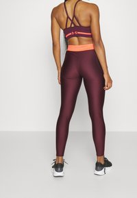 Kobieta w bordowych legginsach z jasnopomarańczowym pasem i pasującym stanikiem sportowym, stojąca plecami do kamery na szarym podłożu.