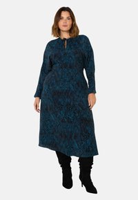 Långärmad klänning i teal och svart mönster med rund halsringning och knytdetalj, åtsmitande vid midjan och utsvängd nertill.