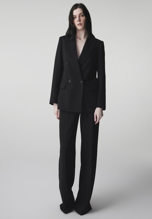 Kobieta o czarnych włosach, ubrana w dopasowaną, czarną, dwurzędową marynarkę i pasujące szerokie spodnie, stojąca na jasnym, jednolitym tle.