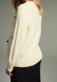 Cream-colored sweater with textured fabric, ribbed hem, and black pom-pom accents. Worn over a fitted black sequined skirt.
