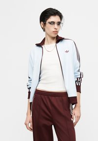 Άτομο που φοράει ανοιχτό μπλε και μπορντό αθλητικό μπουφάν Adidas και μπορντό παντελόνι, λευκή μπλούζα, γυαλιά ηλίου και χρυσή αλυσίδα στο λαιμό, ποζάροντας μπροστά σε λευκό φόντο.