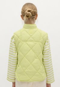 Chaleco acolchado verde lima con cuello alto y detalle de logo en la parte posterior, usado sobre una camisa de manga larga a rayas.