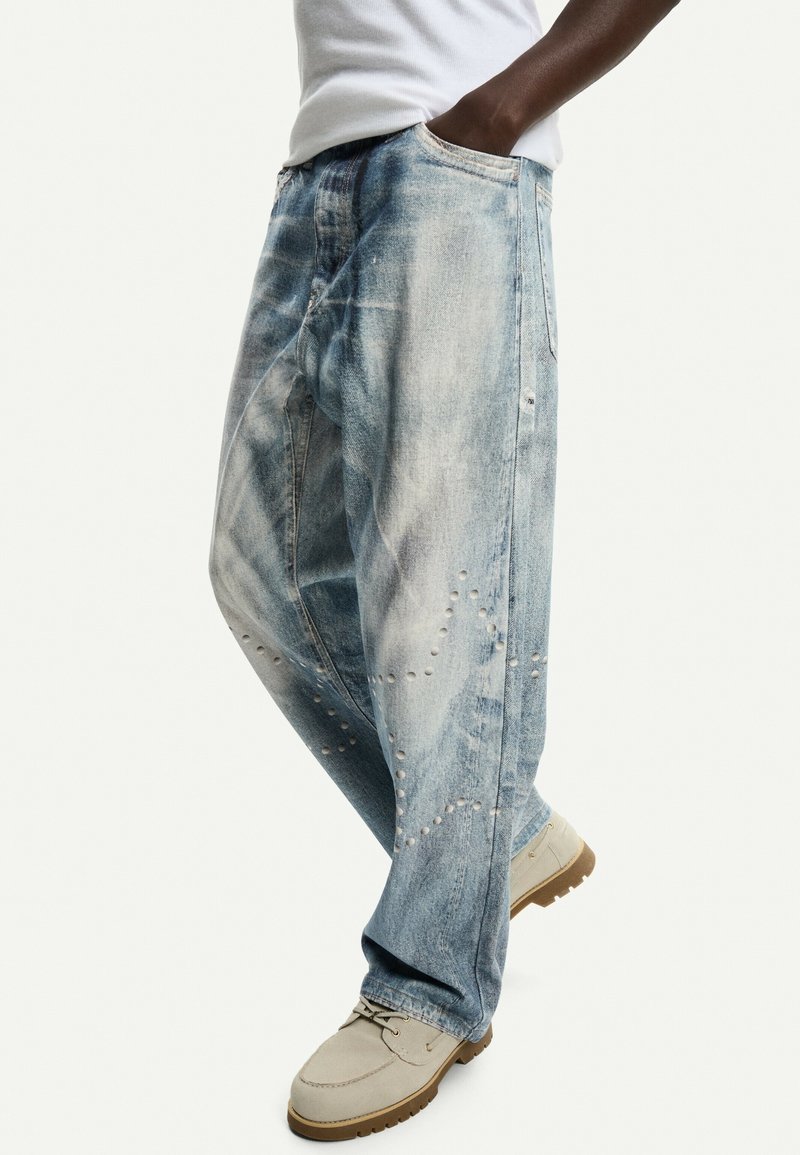 Světlé modré roztrhané džíny s volným střihem, mají vybledlou texturu a dekorativní cvočky po stranách, doplněné béžovými mokasíny.