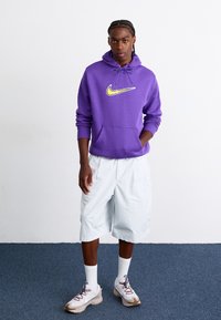 Violetinė Nike gobtuvu su geltona logo, kreminės spalvos sportiniai batai, balti kulkšninių kojinės ir šviesiai mėlynos šortai. Paprastas dizainas su laisvu pritaikymu.