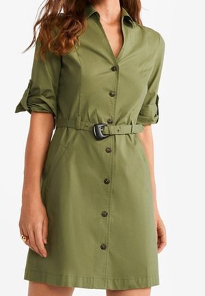 Kvinde iført en kortærmet olivengrøn kjole med knapper foran, matchende bælte og opkrogede ærmer.