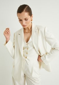 Chaqueta y chaleco de traje a rayas blancas con botones dorados, combinados con pantalones a juego. Tejido texturizado y diseño estructurado. Collar de cadena dorada.