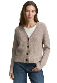 Woman wearing a beige knit buttoned cardigan over a white shirt and blue jeans, standing with one hand in pocket against a white background.