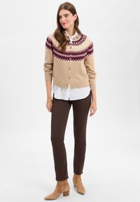 Beige Cardigan mit einer bordeauxfarbenen gestrickten Yoke, die geometrische Muster aufweist, mit Knopfverschluss, kombiniert mit einem weißen Hemd und braunen Hosen. Wildleder-Ankelboots.