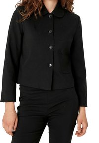 Kobieta ubrana w czarną marynarkę zapinaną na guziki z małą kieszonką po lewej stronie oraz pasującymi czarnymi spodniami, stojąca z relaksującymi się rękami.