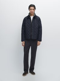 Marineblau gesteppte Jacke mit grauem Kragen, ausgestattet mit Druckknöpfen, kombiniert mit lockeren schwarzen Hosen und braunen Wildlederschuhen. Minimalistisches Design.