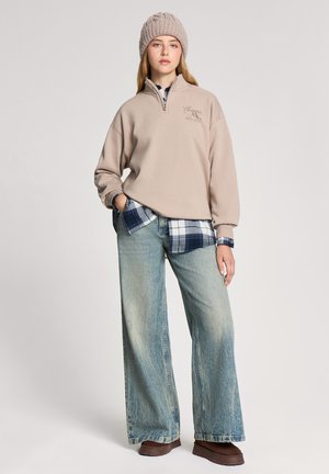 Pullover in pile beige con mezza cerniera, abbinato a una camicia a quadri blu e bianca, jeans a gamba larga sbiaditi e scarpe marroni slip-on.