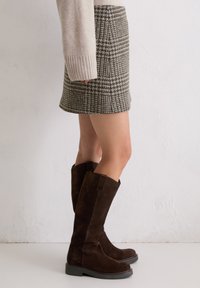 Brązowe zamszowe kozaki do kolan z grubą czarną podeszwą. Projekt charakteryzuje się gładką teksturą i delikatnym, opadającym wykończeniem.